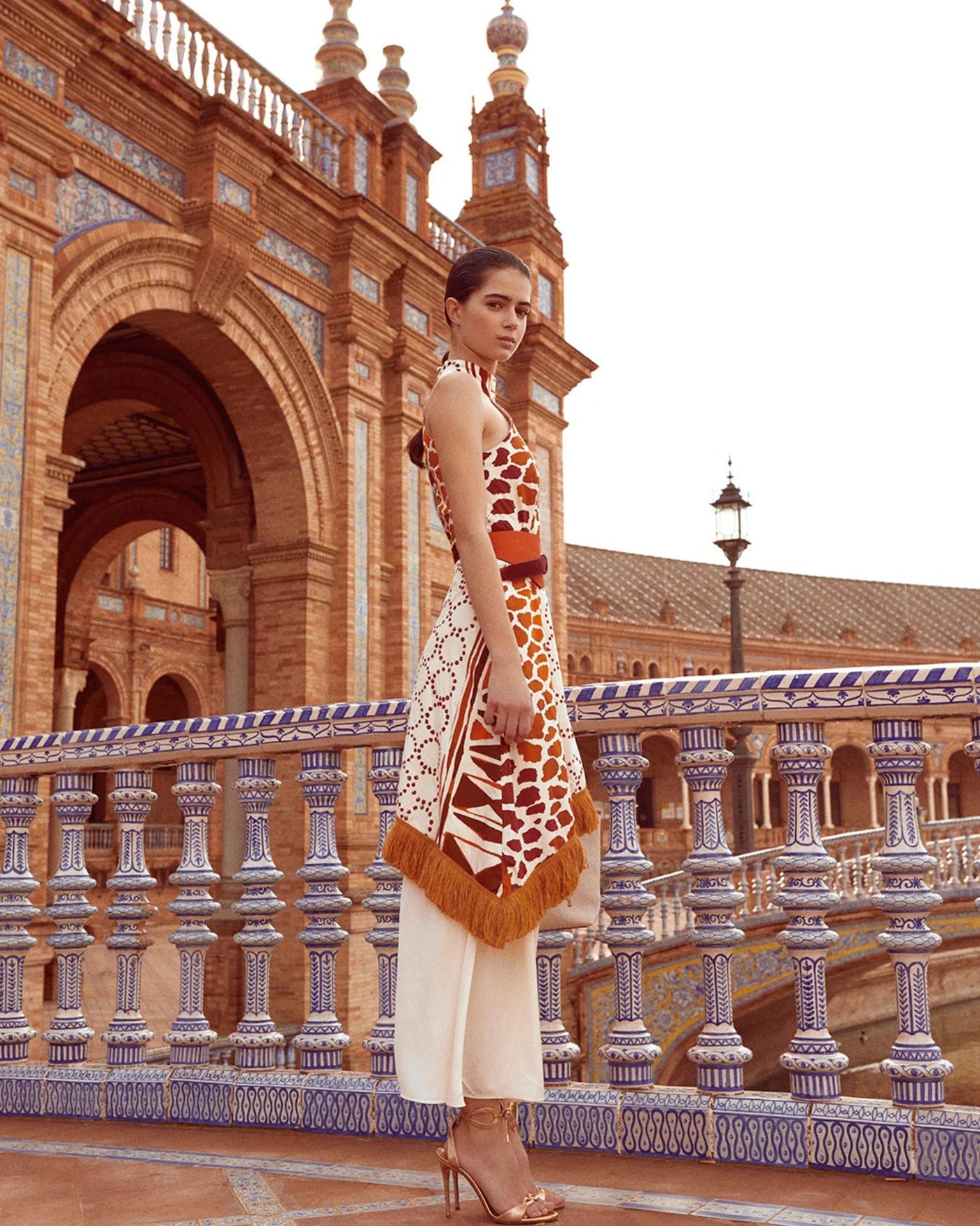 The Eliane Dress is a flowy, long dress with a halter neckline. The ruffled top of the skirt and fringe detailing add a playful touch. It features a sleeveless design and hidden back zipper for a seamless look. The vibrant orange and beige print will make you stand out in any occasion.