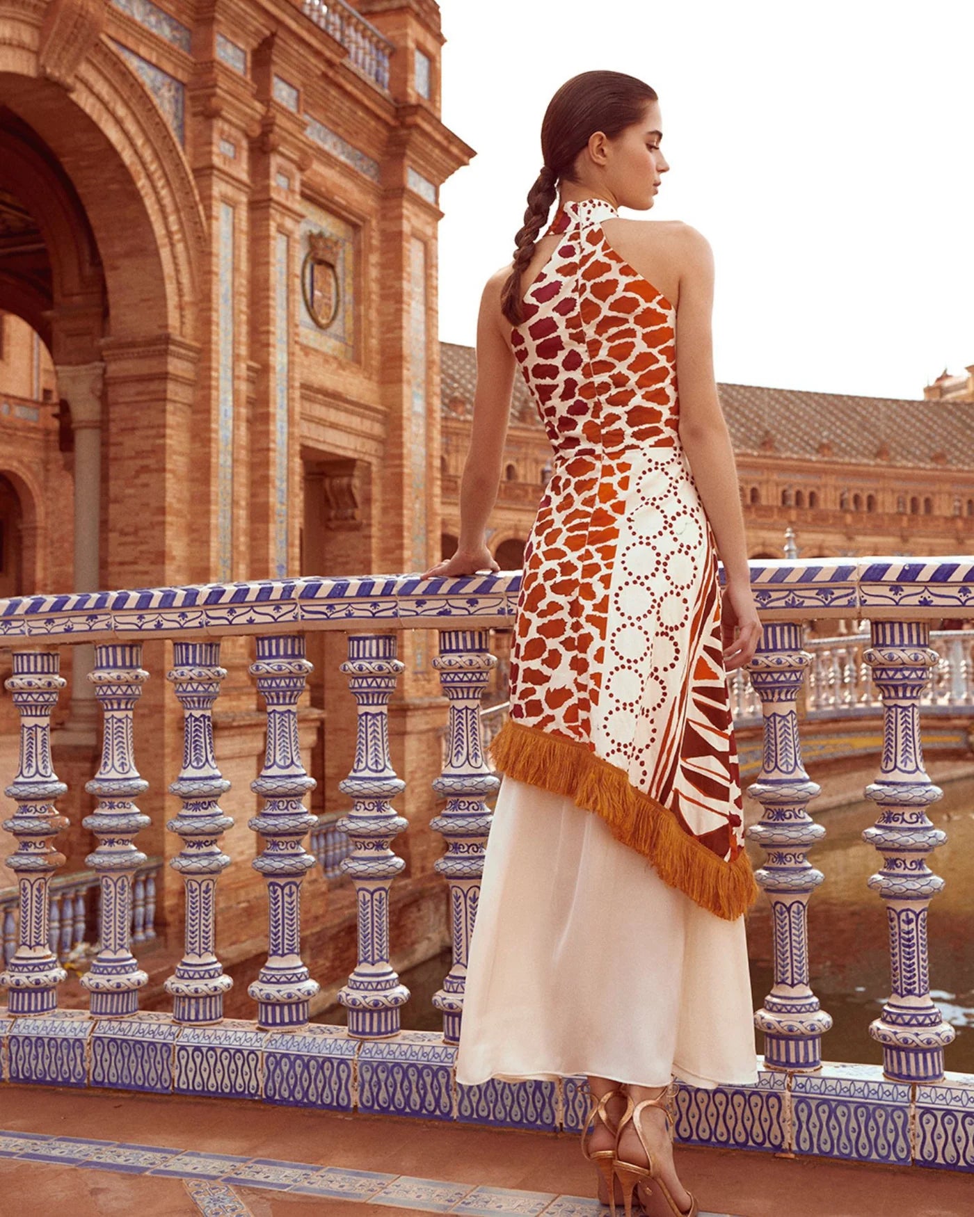 The Eliane Dress is a flowy, long dress with a halter neckline. The ruffled top of the skirt and fringe detailing add a playful touch. It features a sleeveless design and hidden back zipper for a seamless look. The vibrant orange and beige print will make you stand out in any occasion.