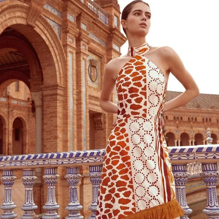 The Eliane Dress is a flowy, long dress with a halter neckline. The ruffled top of the skirt and fringe detailing add a playful touch. It features a sleeveless design and hidden back zipper for a seamless look. The vibrant orange and beige print will make you stand out in any occasion.