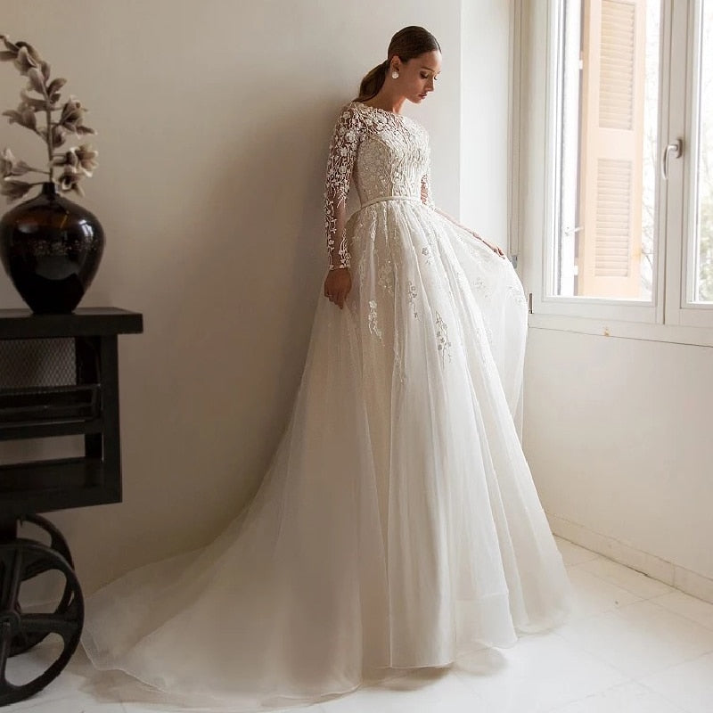 Elevate your wedding look with the Robe Anneliese. This A-line wedding dress features elegant illusion lace long-sleeves and a scoop neckline for a timeless and sophisticated look. The applique tulle train adds a touch of romance and sophistication. Make a statement on your special day in this beautiful bridal gown.