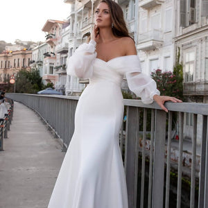 Experience pure elegance with Robe Alora! This bohemian-inspired wedding dress features a sexy satin mermaid silhouette, long lace sleeves, and sultry off-the-shoulder detailing for a ravishing and timeless look. Get ready to turn heads on your special day!